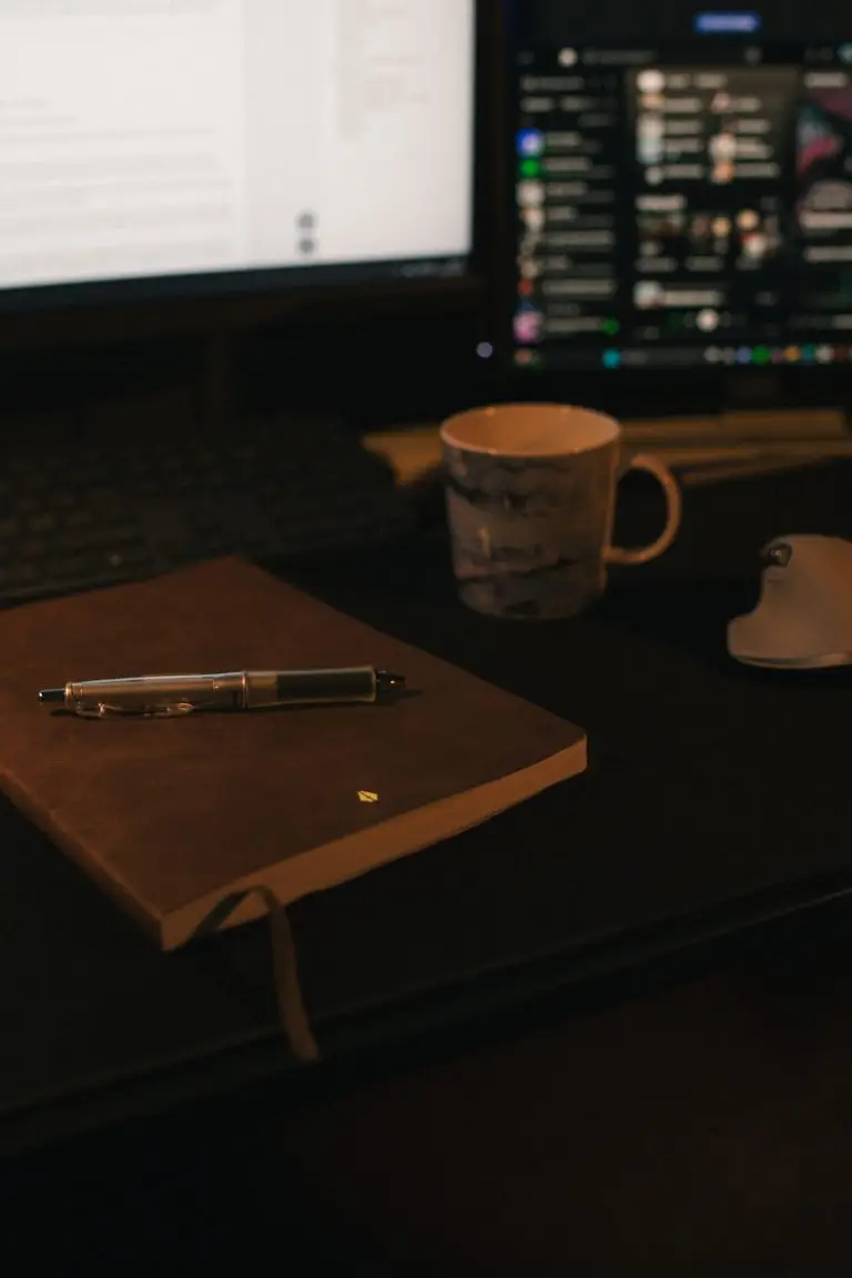 A leather notebook, a pen, and a coffee cup on computer desk